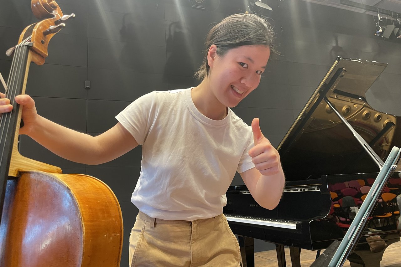 Musikerin mit Cello vor einem Flügel, Daumen nach oben zeigend und lachend