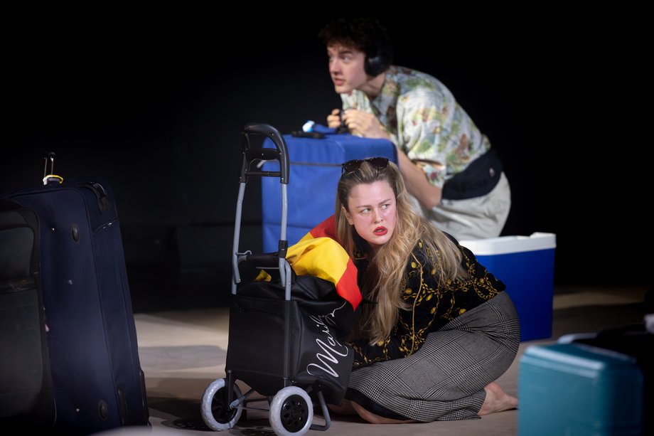 Eine Frau kniet vor einem Einkaufstrolley, hinter ihr ein Mann mit blauem Koffer