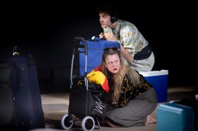 Eine Frau kniet vor einem Einkaufstrolley, hinter ihr ein Mann mit blauem Koffer