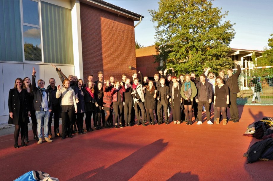 Many people stand in a semicircle in a schoolyard and laugh into the camera